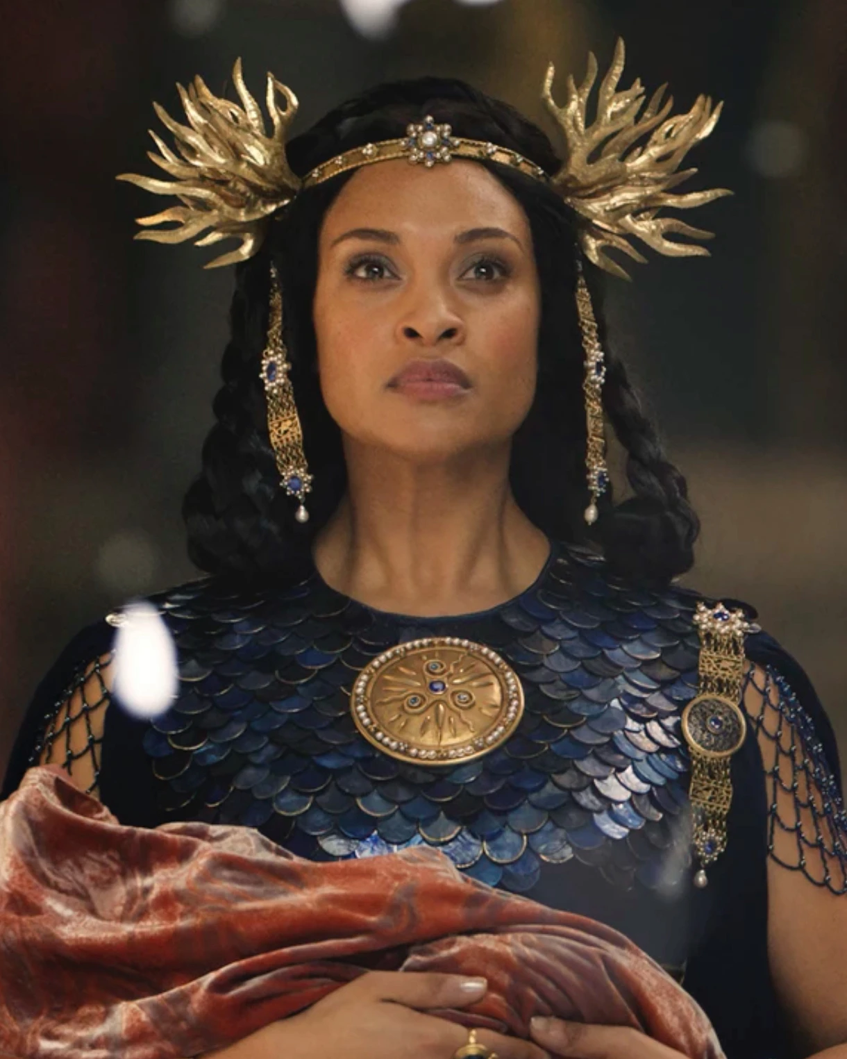 A film character in blue armour wearing a winged gold-coloured crown and a circular brooch.